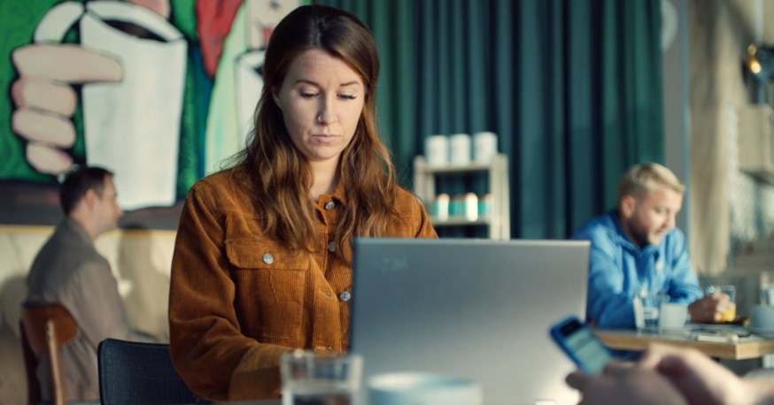 Konsentrert kvinne i brun fløyelsskjorte sitter med laptop på kafé